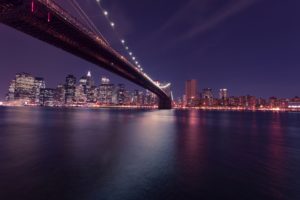 Night view of the Brooklyn Bridge stretching over the East River with the illuminated New York City skyline in the background, reflecting vibrant lights on the water