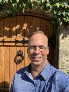 Smiling man wearing glasses and a blue patterned shirt taking a selfie in front of a rustic wooden door framed with ivy and stone
