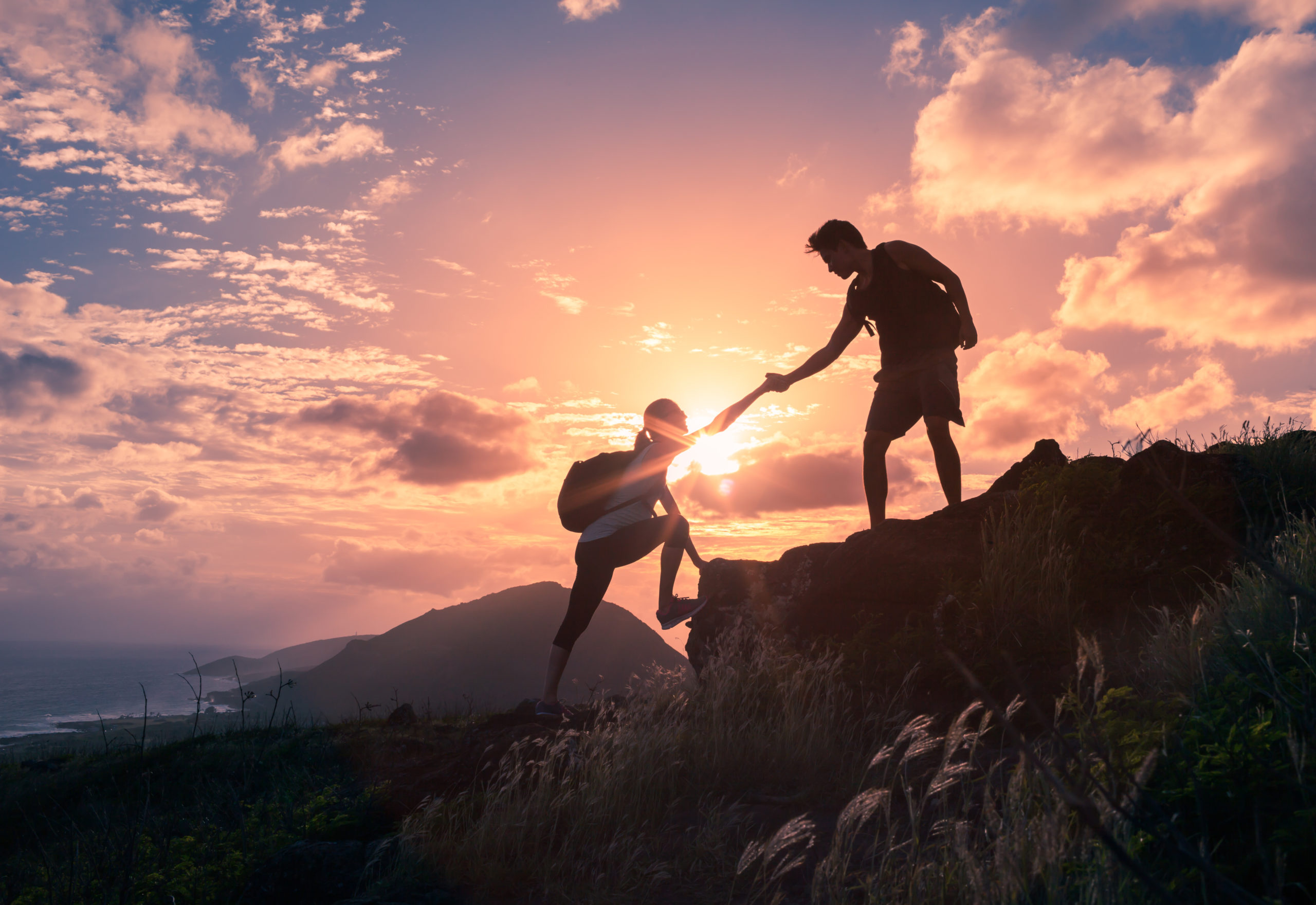 Two hikers silhouetted against a vibrant sunset, with one helping the other climb a rocky hilltop, symbolizing teamwork, support, and adventure in nature