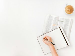 Flat lay of a hand writing in a journal next to an open Bible and a cup of coffee on a white background, symbolizing Bible study, reflection, and quiet time with God