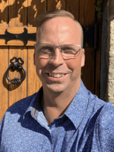 Smiling man wearing glasses and a blue patterned shirt taking a selfie in front of a rustic wooden door framed with ivy and stone