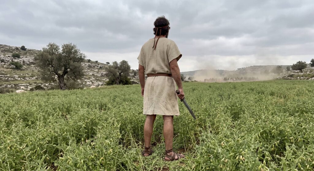 An Israelite man in a wool tunic stands with a sword in the middle of a lentil field as dust rises from an approaching army in the hills