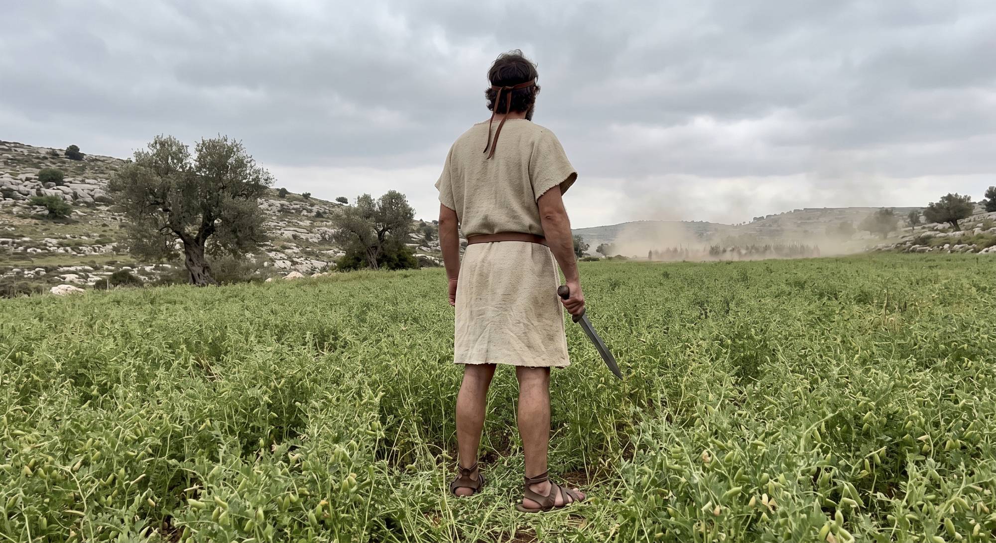 An Israelite man in a wool tunic stands with a sword in the middle of a lentil field as dust rises from an approaching army in the hills